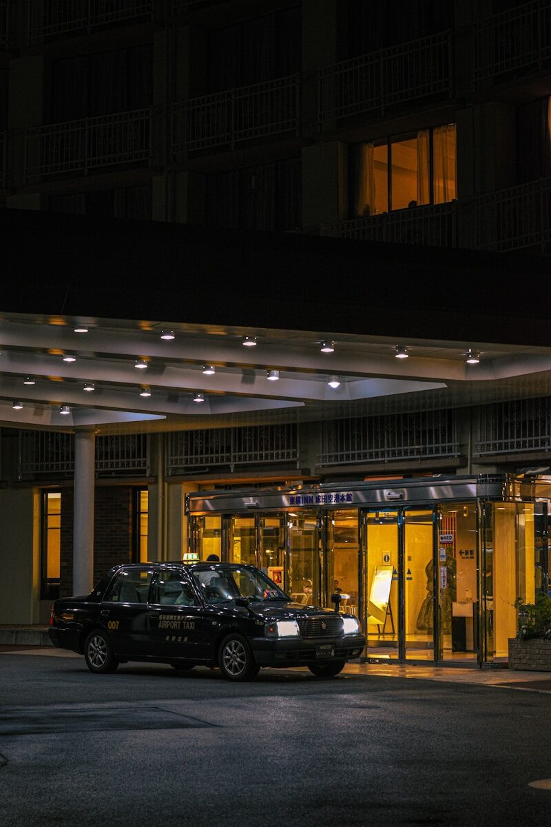 A black taxi waits outside a brightly lit hotel entrance.