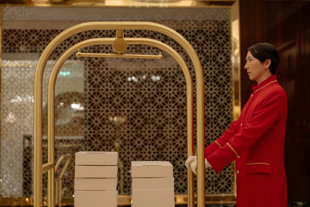 A hotel bellboy in uniform pushes a luggage cart with boxes indoors, demonstrating hospitality service.