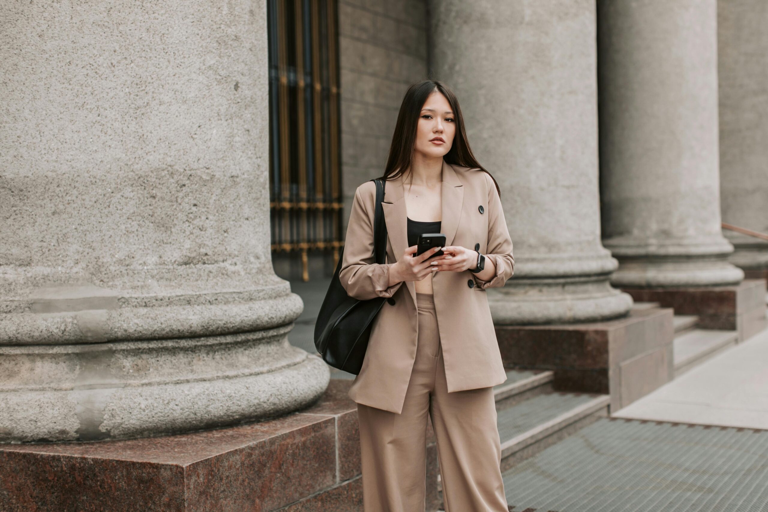 Young woman in beige business suit holding smartphone, standing near columns outside.