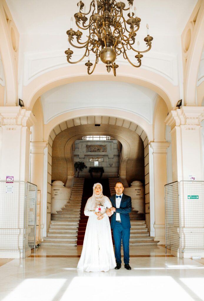 A bride and groom pose elegantly beneath a grand archway with intricate architecture.